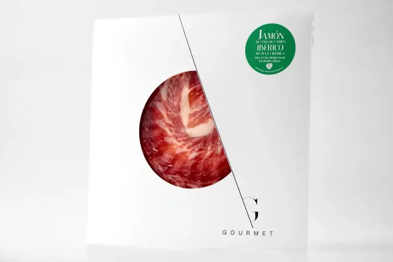 Paquete de Guijuelo Gourmet Jamón de Cebo de Campo 50% Ibérica, con un diseño de sobre blanco cuadrado, presenta un corte circular en el centro que permite ver el producto del jamón, y lleva una etiqueta circular verde en la esquina superior derecha.