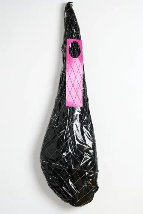 Bolsa de tela blanca con la pierna entera de jamón de cebo 50% ibérica de Guijuelo Gourmet, envuelta en una red blanca poco densa, con una etiqueta de papel de color rosa y el logotipo en negro impreso en la tela.