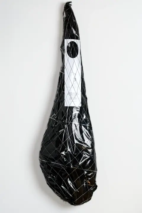 Jamón ibérico de pata entera en bolsa verde, envuelto en una red blanca y con etiqueta de papel blanco; el logotipo en blanco está impreso en la bolsa.