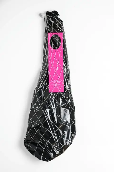 Bolsa de tela blanca con la pierna entera de paleta de cebo 50% ibérica de Guijuelo Gourmet, envuelta en una red blanca poco densa, con una etiqueta de papel de color rosa y el logotipo en negro impreso en la tela.
