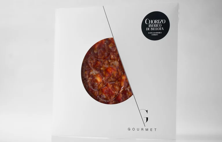 Paquete blanco cuadrado con un recorte circular en el centro que permite ver el chorizo ibérico, con una etiqueta negra en la esquina superior derecha.